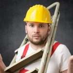 A confused handyman stands with a step ladder around his neck while holding a hammer